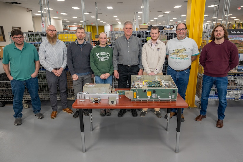 Eight people pose for a photo behind a table with pieces of a Power Amplifier Module, a piece of military equipment on it.