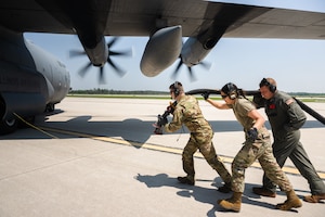 three airmen pull a fuel hose to an aircraft with moving propellers