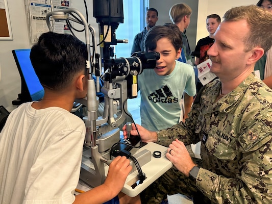 Lt. Trey Ketchum, an optometrist at U.S. Naval Hospital Guantanamo Bay, demonstrates the use of a slit lamp biomicroscope to students from W.T. Sampson Elementary and High School during a field trip, March 10, 2026. Student Noah Calvo looks through the biomicroscope to examine a classmate’s eye as part of the hands-on learning experience. The visit was part of an educational outreach initiative by U.S. Navy Medicine Readiness and Training Command Guantanamo Bay to introduce students to careers in military medicine. (U.S. Navy photo by Emily McCamy)