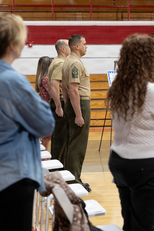 U.S. Marine Corps Sgt. Maj. Joseph P. Olander, center, senior enlisted leader, and Lt. Col. James P. Psyhogis, commanding officer, both with Center for Naval Aviation Technical Training Unit New River, stand at attention during the U.S. National Anthem during an Adopt-a-School ceremony at Jacksonville High School in Jacksonville, North Carolina, March 18, 2026. The final adoption ceremony between Jacksonville High School and the Center for Naval Aviation Technical Training Unit New River marked a significant achievement for Onslow County School District as it reached a 100 percent adoption rate by various units of Marine Corps Installations East and II Marine Expeditionary Force in the Adopt-a-School program. This initiative aims to strengthen the bond between the military and the local community. (U.S. Marine Corps photo by Lance Cpl. Alyssa J. DeCrane)