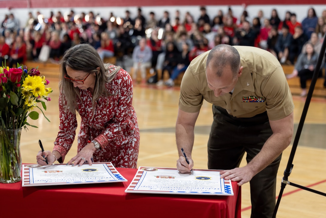 U.S. Marine Corps Lt. Col. James P. Psyhogis, right, commanding officer, Center for Naval Aviation Technical Training Unit New River, and Brenda Hermann, the principal of Jacksonville High School, sign the adoption proclamation during an Adopt-a-School ceremony at Jacksonville High School in Jacksonville, North Carolina, March 18, 2026. The final adoption ceremony between Jacksonville High School and the Center for Naval Aviation Technical Training Unit New River marked a significant achievement for Onslow County School District as it reached a 100 percent adoption rate by various units of Marine Corps Installations East and II Marine Expeditionary Force in the Adopt-a-School program. This initiative aims to strengthen the bond between the military and the local community. (U.S. Marine Corps photo by Lance Cpl. Alyssa J. DeCrane)