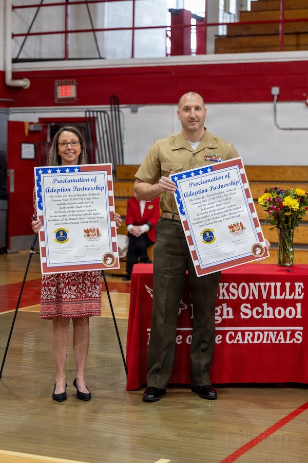 U.S. Marine Corps Lt. Col. James P. Psyhogis, right, commanding officer, Center for Naval Aviation Technical Training Unit New River, and Brenda Hermann, the principal of Jacksonville High School, pose for a photo with the adoption proclamations during an Adopt-a-School ceremony at Jacksonville High School in Jacksonville, North Carolina, March 18, 2026. The final adoption ceremony between Jacksonville High School and the Center for Naval Aviation Technical Training Unit New River marked a significant achievement for Onslow County School District as it reached a 100 percent adoption rate by various units of Marine Corps Installations East and II Marine Expeditionary Force in the Adopt-a-School program. This initiative aims to strengthen the bond between the military and the local community. (U.S. Marine Corps photo by Lance Cpl. Alyssa J. DeCrane)