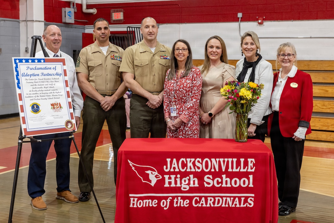 U.S. Marine Corps Center for Naval Aviation Technical Training Unit New River leaders, Marine Corps Community Services School Liaisons, and Jacksonville High School faculty, pose for a photo during the U.S. National Anthem during an Adopt-a-School ceremony at Jacksonville High School in Jacksonville, North Carolina, March 18, 2026. The final adoption ceremony between Jacksonville High School and the Center for Naval Aviation Technical Training Unit New River marked a significant achievement for Onslow County School District as it reached a 100 percent adoption rate by various units of Marine Corps Installations East and II Marine Expeditionary Force in the Adopt-a-School program. This initiative aims to strengthen the bond between the military and the local community. (U.S. Marine Corps photo by Lance Cpl. Alyssa J. DeCrane)