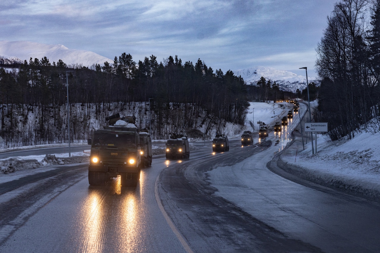 A convoy of military vehicles drives down a road in snow-covered hills with mountains in the background.