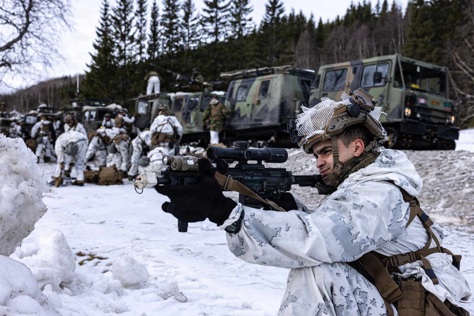 A man in winter camouflage gear kneels in the snow while aiming his rifle. A group of people in similar attire check their gear in the background.