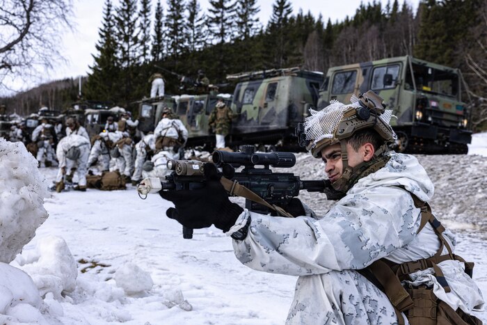 A man in a winter camouflage military uniform kneels in the snow while aiming his rifle. A group of people in similar attire check their gear in the background. There are also military vehicles parked on a road behind them.