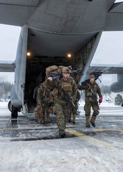 People in camouflage military uniforms exit the back of a large military aircraft on a tarmac.