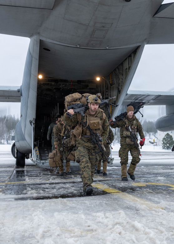 People in camouflage military uniforms exit the back of a large military aircraft on a tarmac.