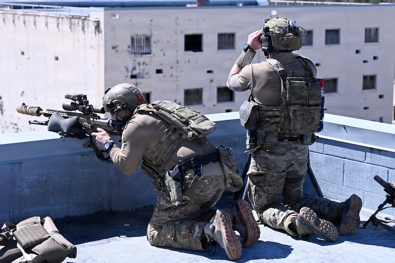 Two men wearing camouflage military uniforms and tactical helmets with hearing protection are on a rooftop; one aims a rifle at a target while the other looks through binoculars.