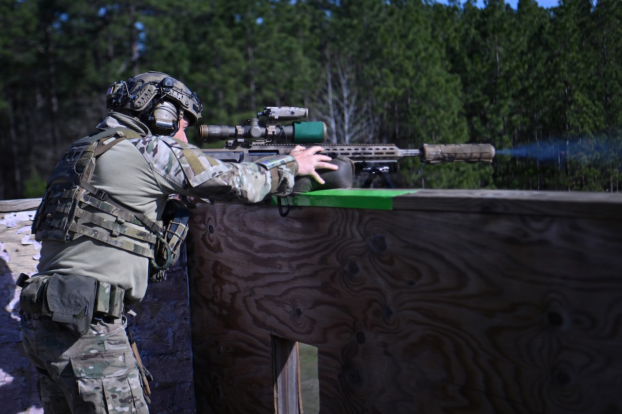 A man wearing a military camouflage uniform and a tactical helmet with hearing protection aims and fires a rifle with the support of a plywood wall.