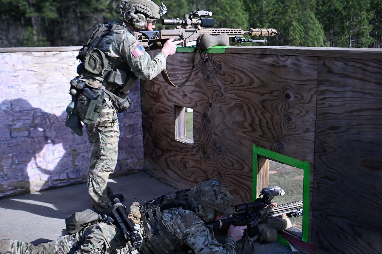 Two men wearing camouflage military uniforms and tactical helmets with hearing protection point rifles at targets. One man is standing, using a plywood wall as a support for his rifle, while the other is lying in a prone position, aiming his weapon through a hole in the wall.