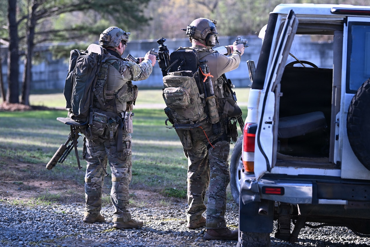 Two men wearing camouflage military uniforms, backpacks and tactical helmets with hearing protection stand next to a vehicle while aiming pistols at a target.