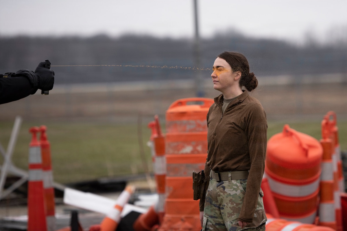A hand from the left sprays orange oleoresin capsicum on an airman's eyes while standing next to traffic cones on a gloomy day.