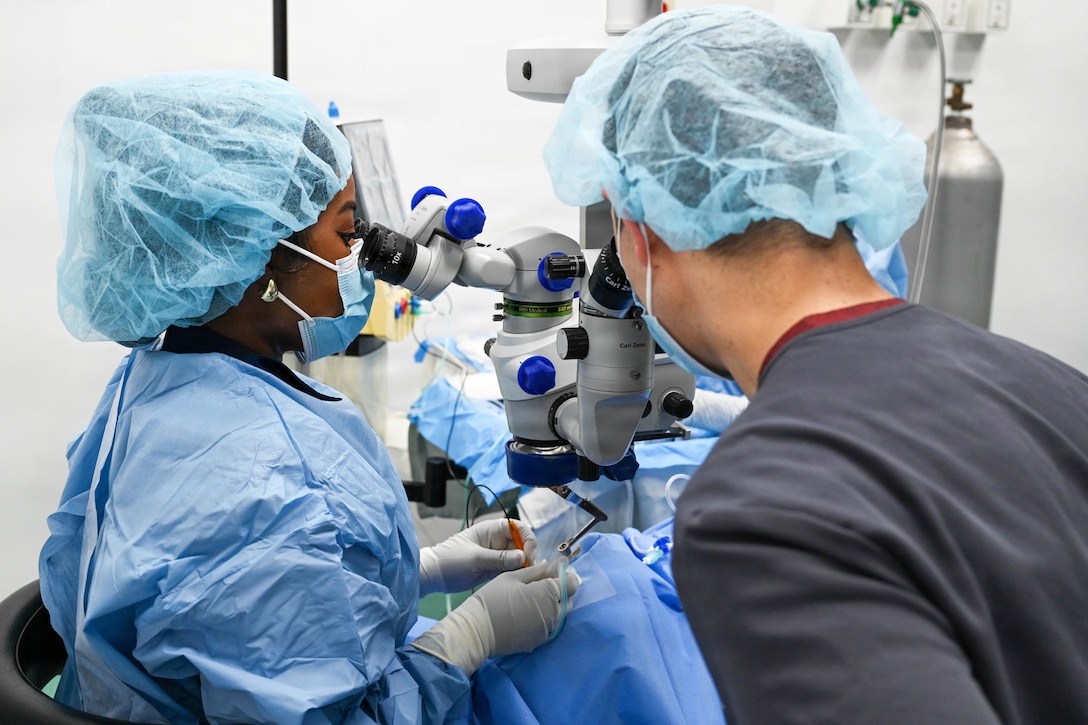 A surgeon in protective gear looks through a tool to perform a surgery as a fellow doctor advises.