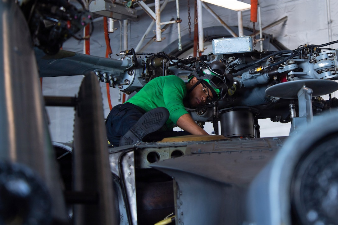 A sailor in a green helmet and shirt works on top of a helicopter in a hangar bay of a ship.