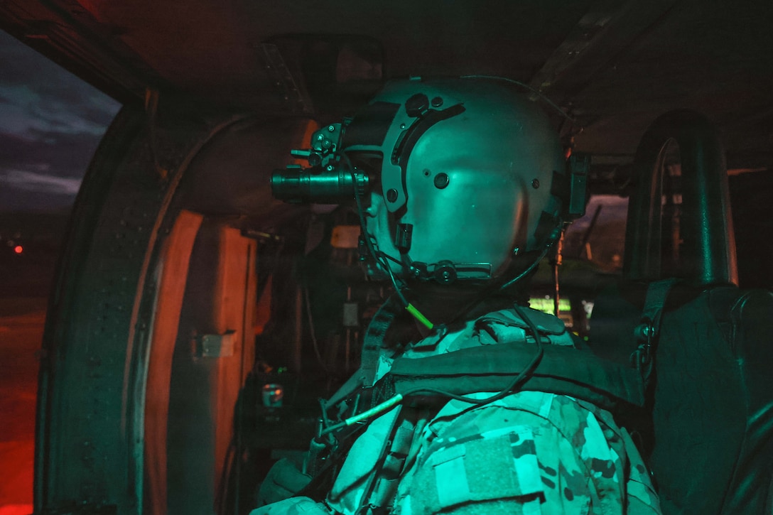 The side view of a soldier in tactical gear using binoculars while sitting in a helicopter in the dark.