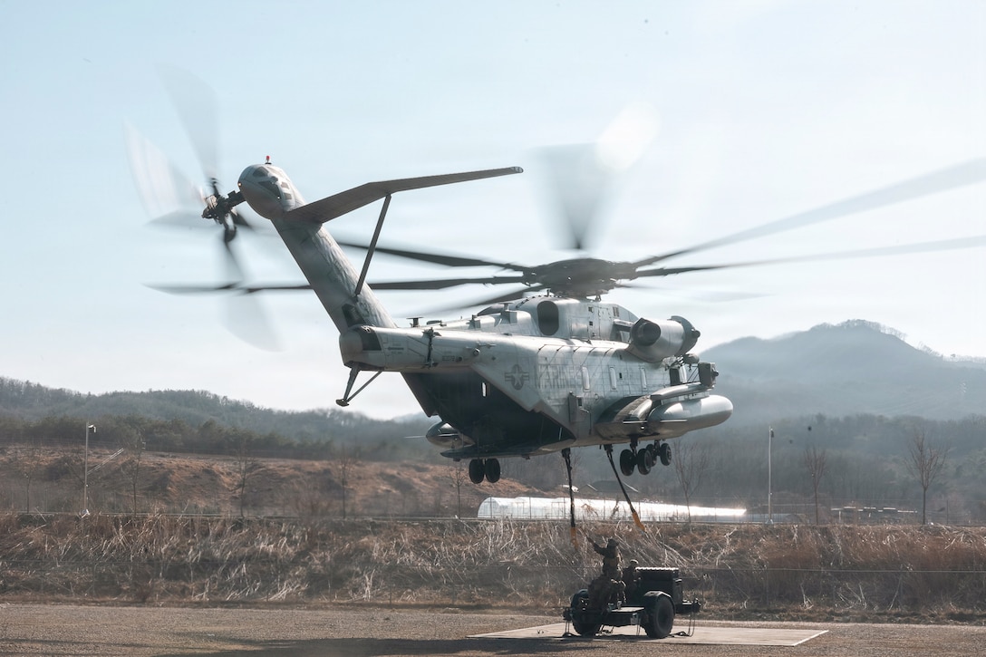 Marines prepare to attach a trailer to a hovering helicopter on a paved road with mountains in the background.