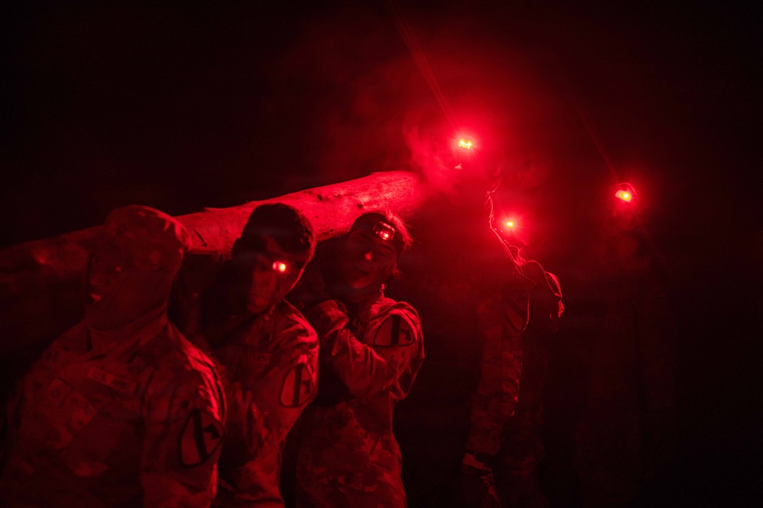 Soldiers carry a log in the dark illuminated by red headlamps.
