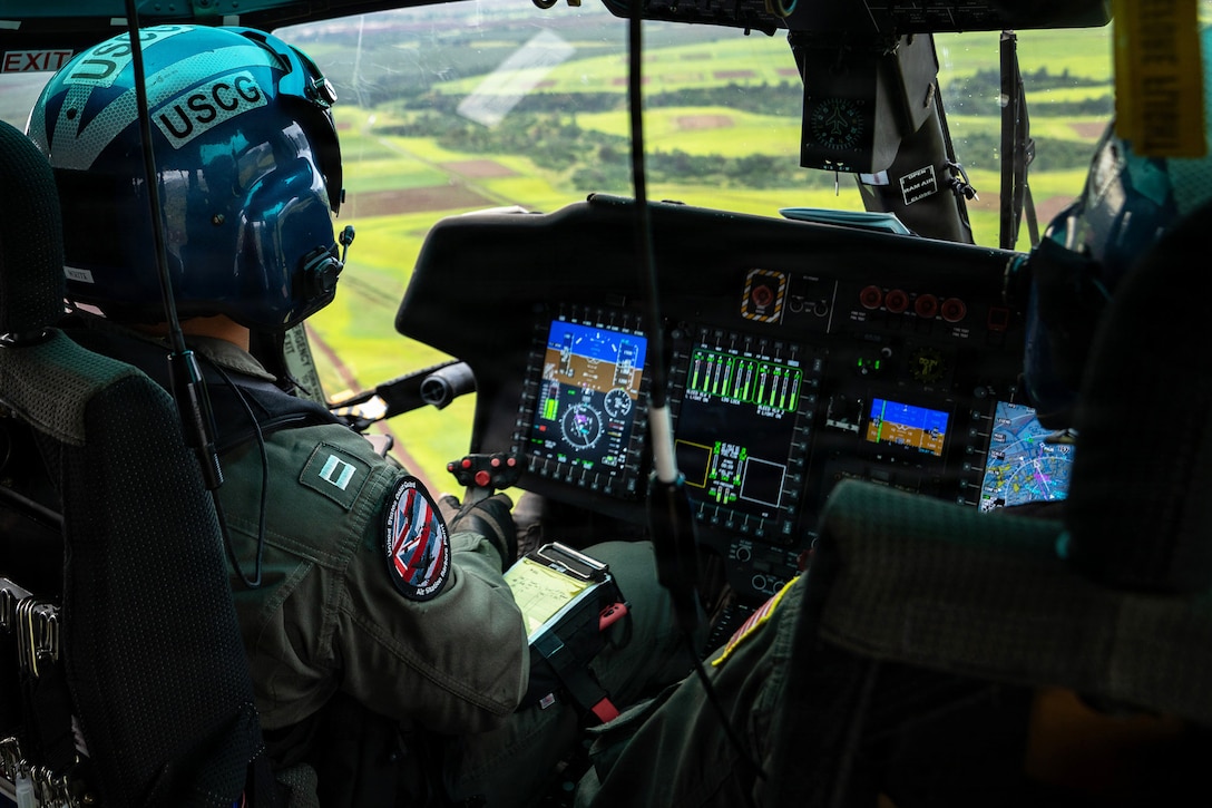 A Coast Guardsman sits in the cockpit of a helicopter as it flies over a green field.