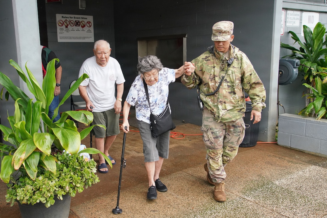A National Guardsman helps support an elderly person walking with a cane outside a building.