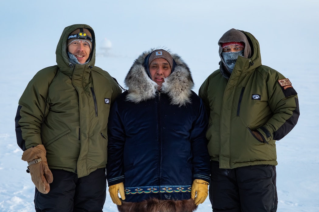 Three men in cold weather attire pose for a group photo out in the frozen tundra.