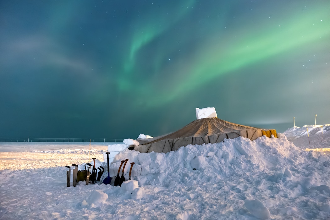 A tent sits buried amongst deep snow in the frozen tundra while the northern lights dance across the sky.