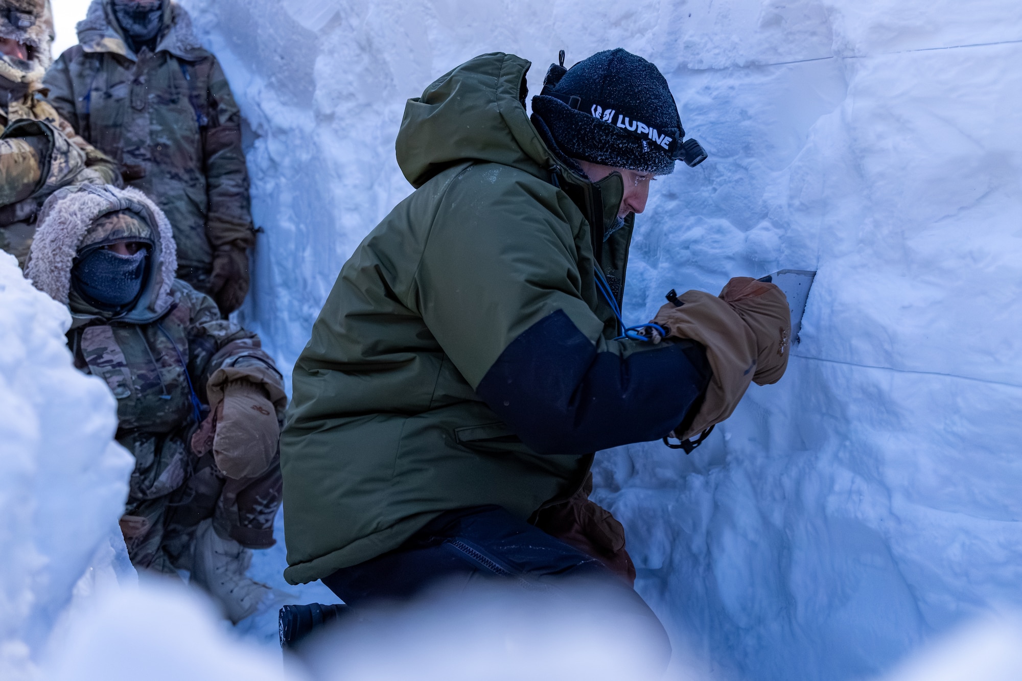 Man kneeling in deep snow cuts through more snow using tool.