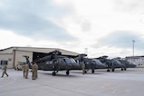four military helicopters in a line and people wearing military uniforms walking around