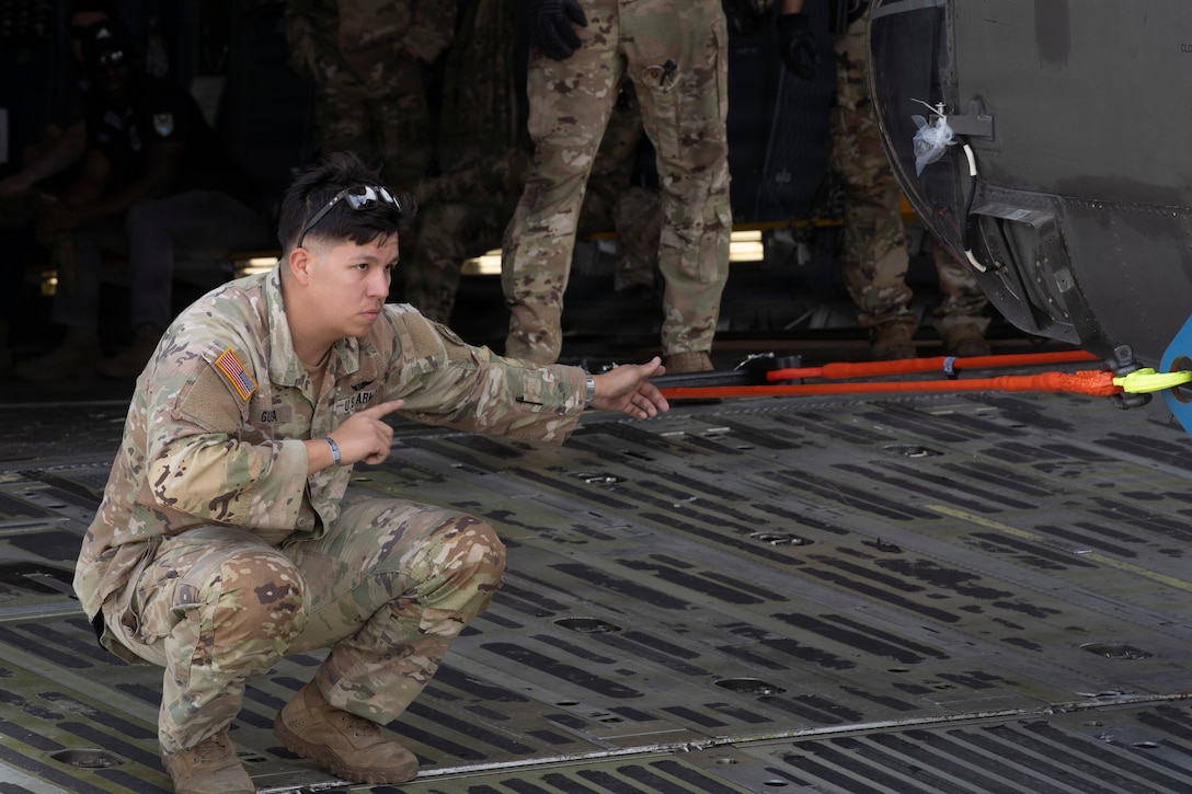 A military member stands on a military plane and guides a helicopter.