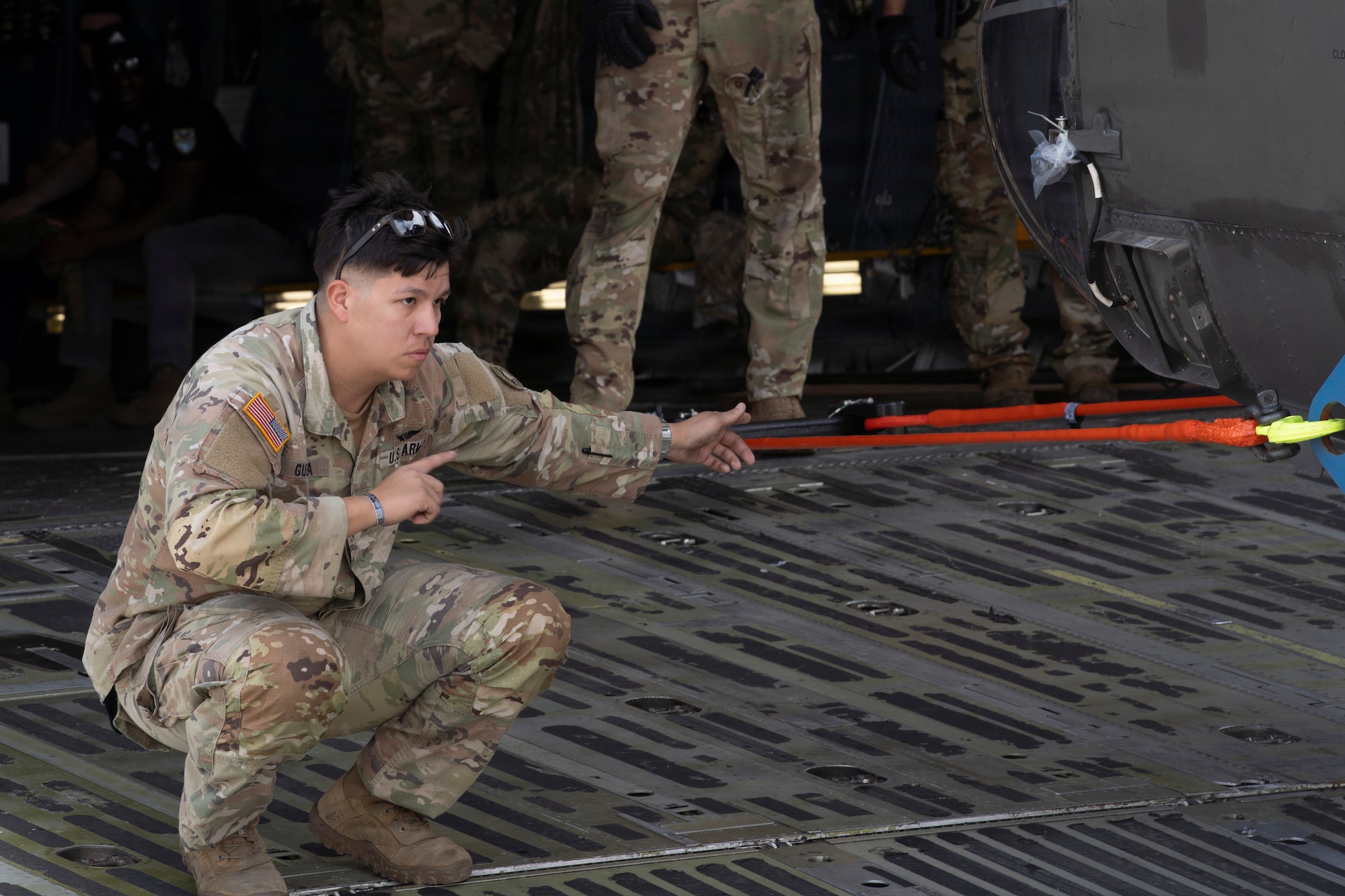 A military member stands on a military plane and guides a helicopter.