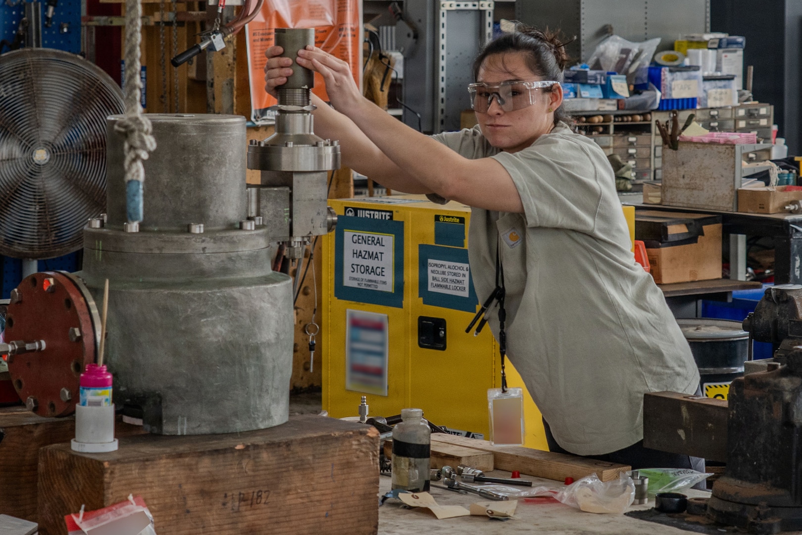 A brown-haired woman reaches up to a shiny and tall master cylinder apparatus to test a part before installing it. She is on board a ship and is in a maintenance shop.