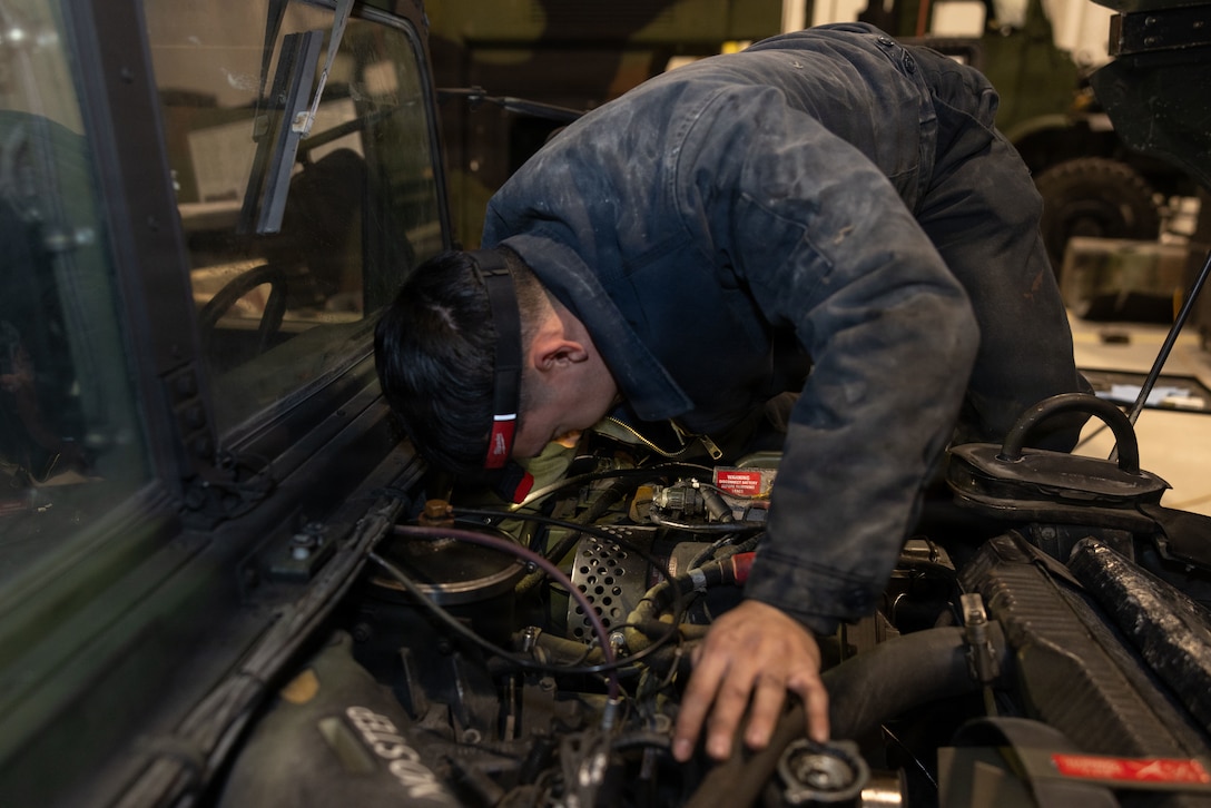 A dark haired man in a blue jumpsuit sits in the engine compartment of a vehicle reaching down into the compartment.