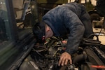 A dark haired man in a blue jumpsuit sits in the engine compartment of a vehicle reaching down into the compartment.