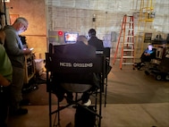 A chair with NCIS:Origins printed on it is viewed from behind while crew from the show monitor a scene from the screen in the background.