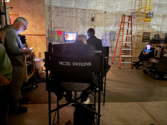 A chair with NCIS:Origins printed on it is viewed from behind while crew from the show monitor a scene from the screen in the background.
