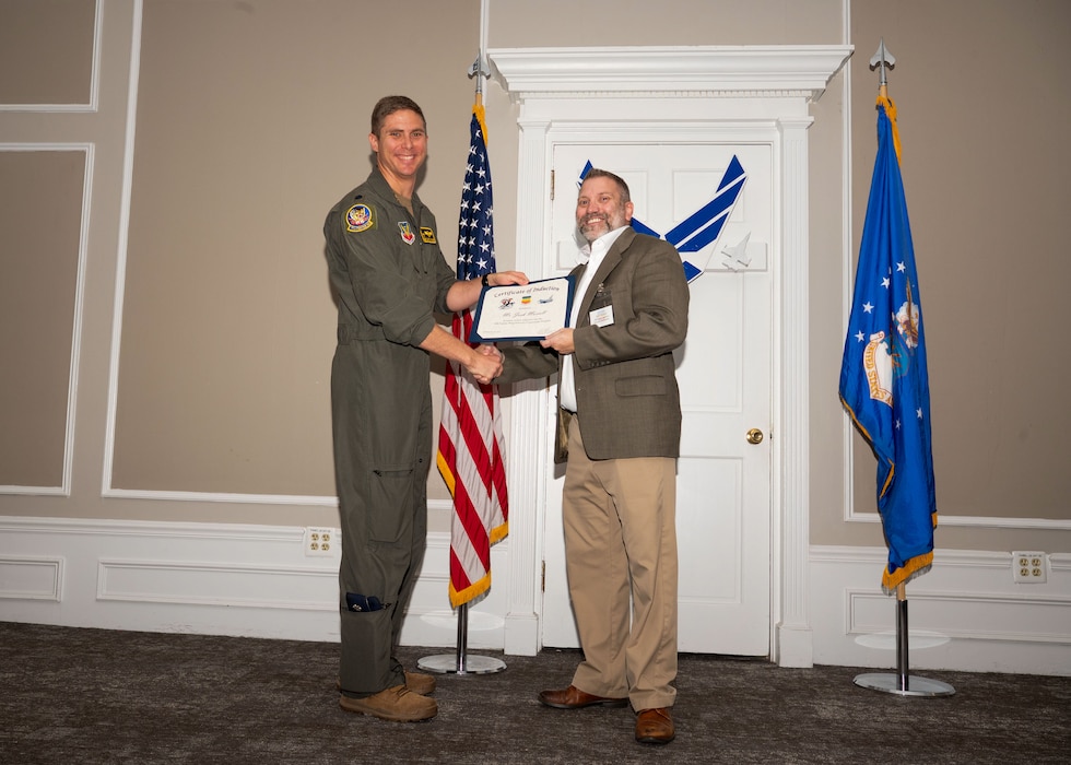 Service member and Honorary Commander shake hands while holding a certificate.