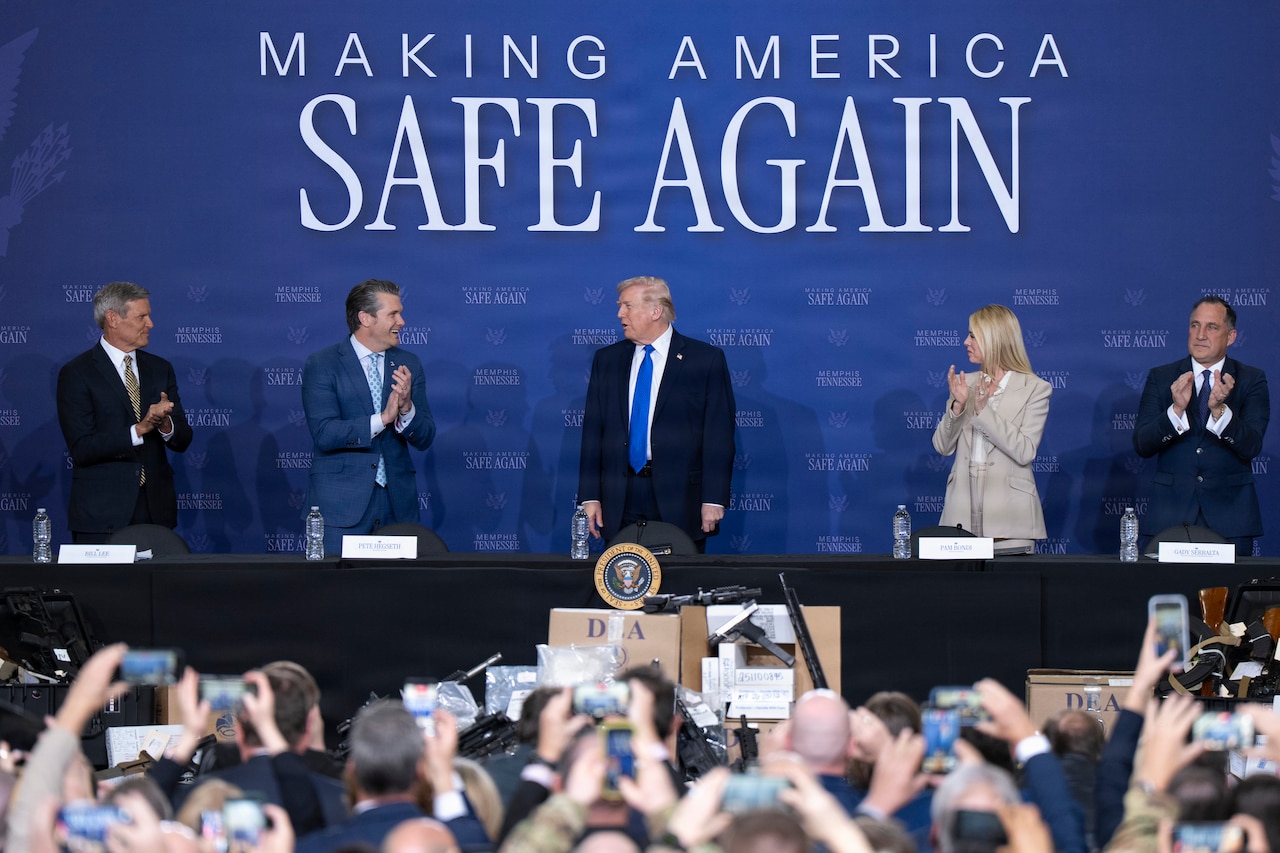 The secretary of war and three other leaders applaud the president as they stand at a table in front of a blue background.