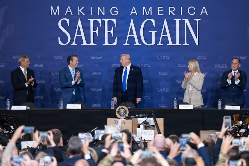 The secretary of war and three other leaders applaud the president as they stand at a table in front of a blue background.