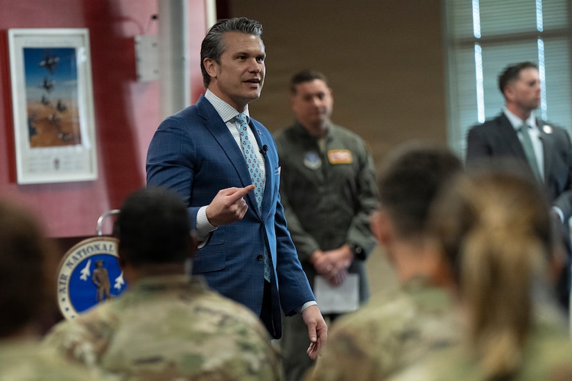 A man in a business suit stands in a room, gesturing with his right hand as he speaks to men and women in camouflage military uniforms with their backs to the camera.