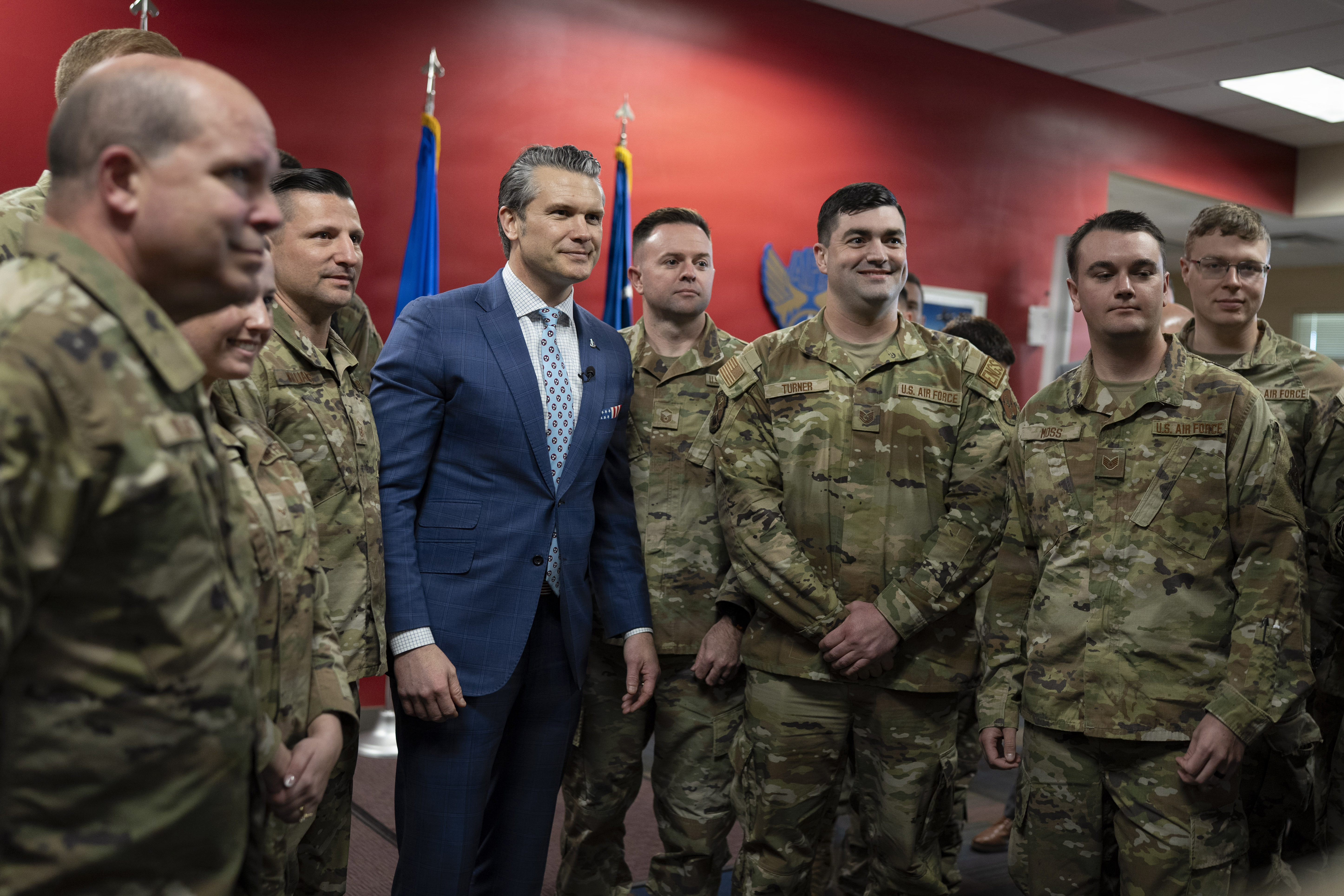 A man in a blue suit poses with troops for a photo in a red-painted room indoors.
