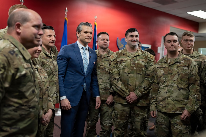 A man in a blue suit poses with troops for a photo in a red-painted room indoors.