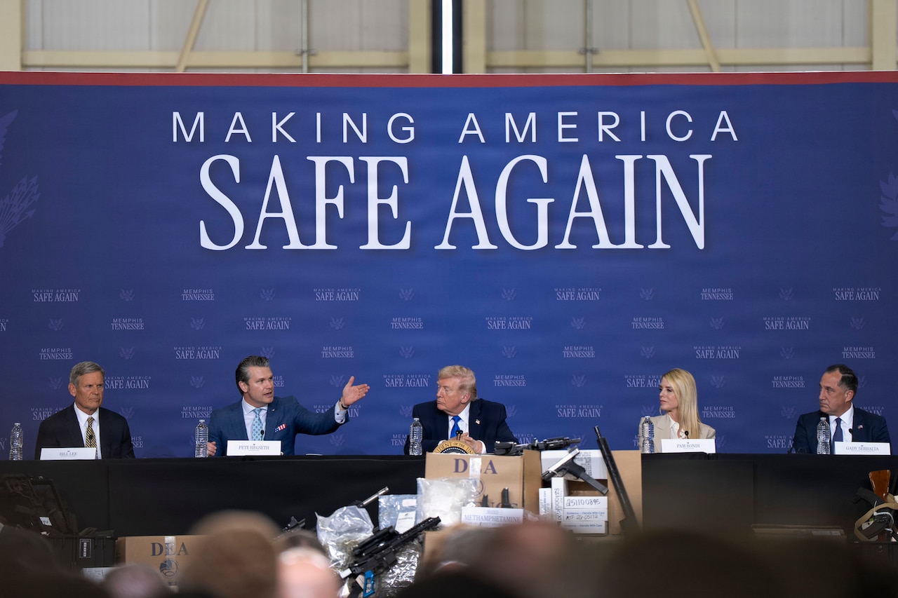 Four men and one woman, all wearing business attire, sit in a row at a long table on a stage. Behind them is a large sign that reads, "Making America Safe Again." One of the men is talking and gesturing with his hand.