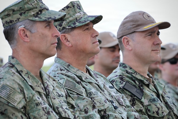 Three military leaders sit and attentively listen to a speech in uniform.