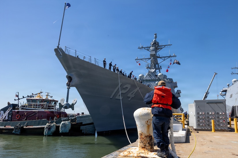 A person in a military uniform and life jacket ties the mooring line of a large military ship to a pier, as several people in military uniforms stand on the bow.