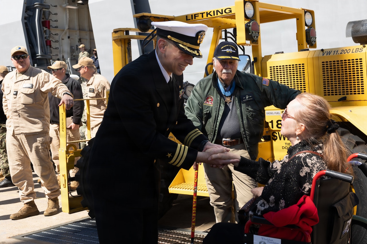 A man in a military dress uniform shakes hands with a woman in a wheelchair while standing on a pier; an elderly man in civilian attire and four men in military uniforms stand in the background.