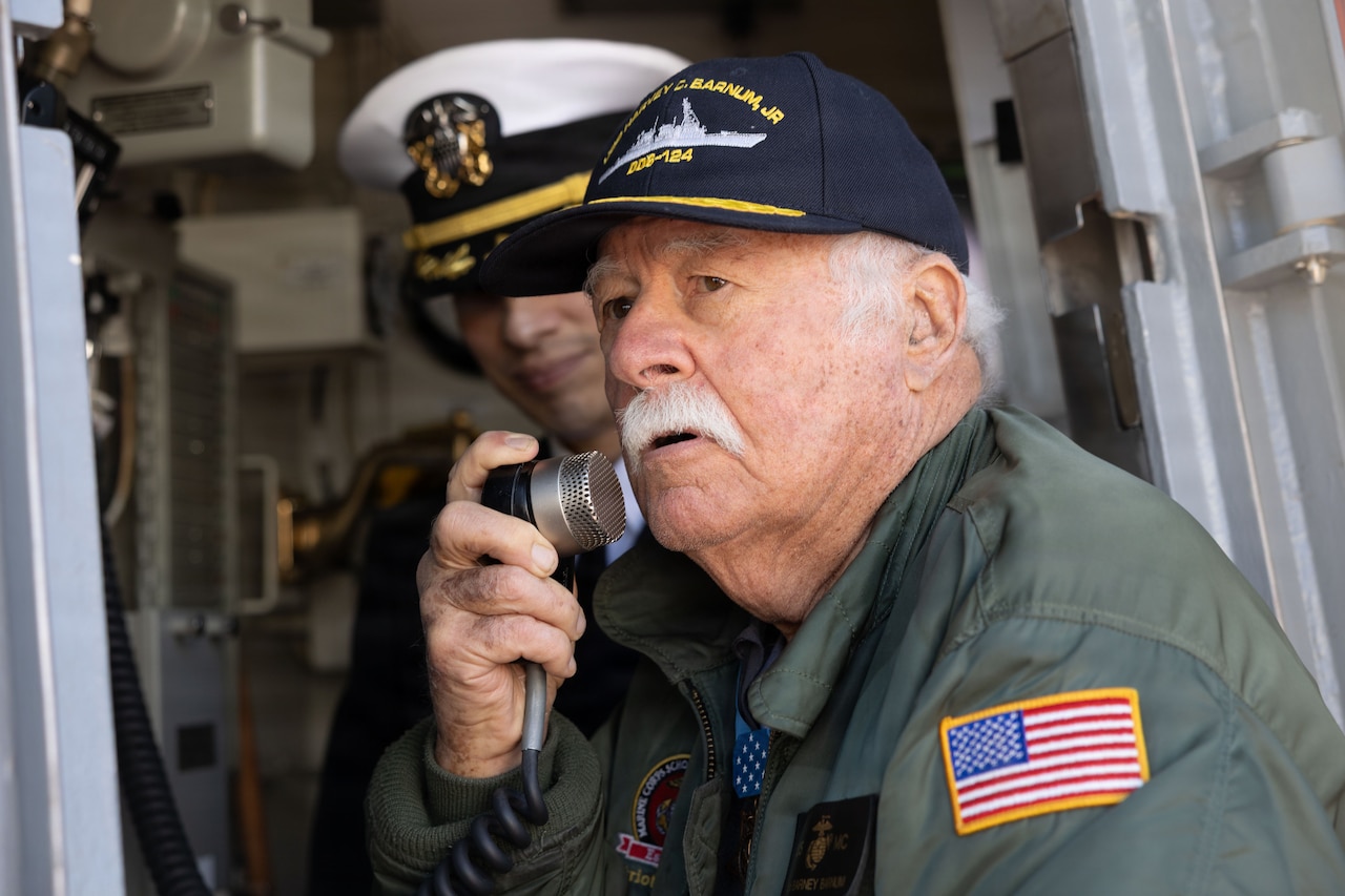 An elderly man speaks into a microphone while on a large military ship. The man is wearing a jacket with an American flag sewn on the shoulder and a command baseball hat with the ship's name on it; another man in a military dress uniform stands in the background.
