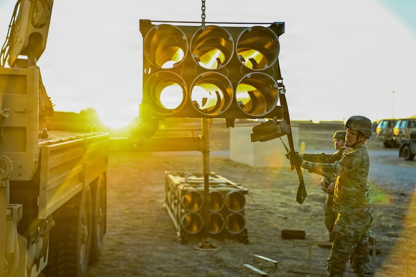 Two soldiers dressed in camouflage military uniforms lift a box of metal tubes using a military vehicle crane.