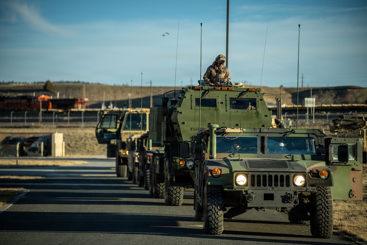 A soldier dressed in a camouflage military uniform stands in the opening of a military vehicle as it drives down a road in a formation with other military vehicles.