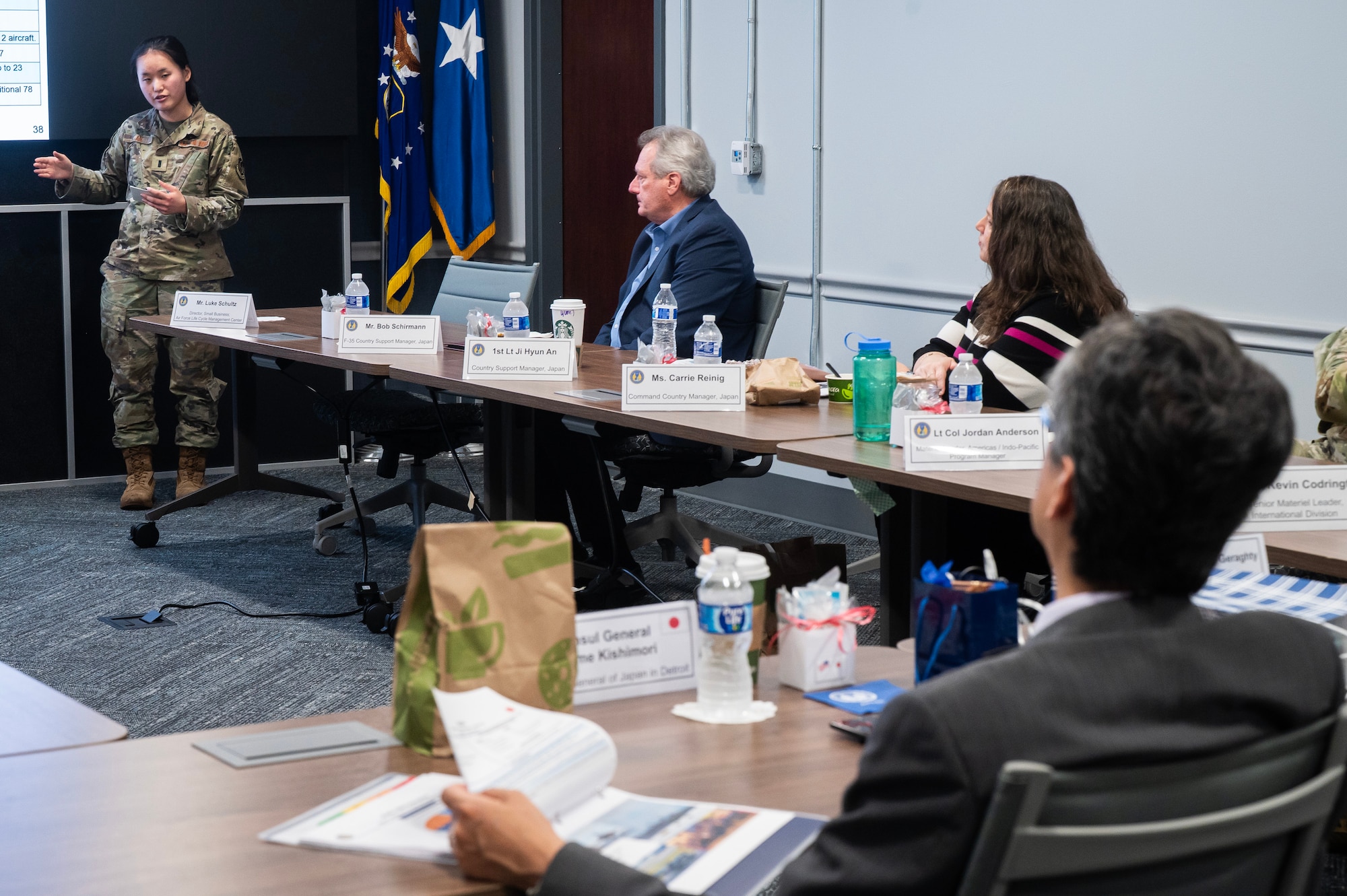 Air Force lieutenant stands at screen and briefs individuals sitting at a table.