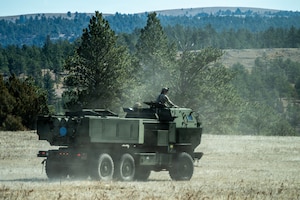 A soldier dressed in a camouflage military uniform stands in the opening of a military vehicle as it drives onto a grassy field.